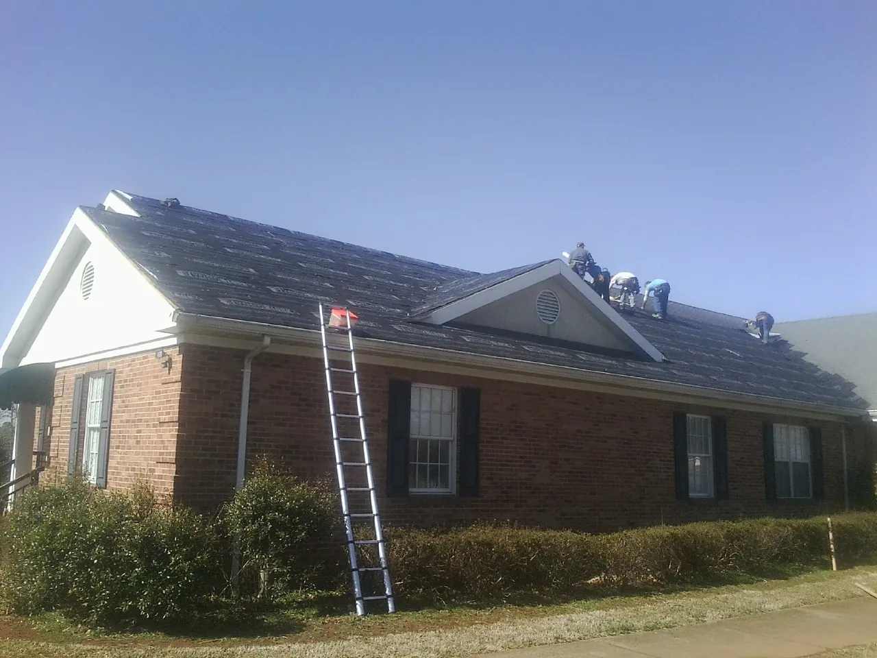 Crew tearing shingles off a brick ranch roof with a ladder set — Emergency Roof Repair work by our crew in New Albin