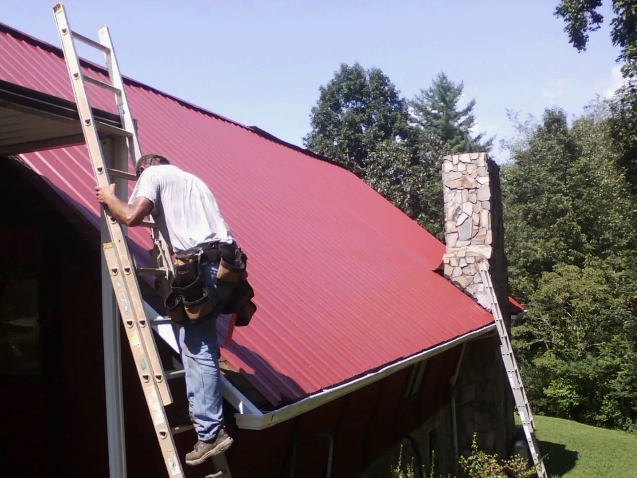 Roofer on a ladder beside a red metal roof and stone chimney — Soffit & Fascia Repair by Roofing Brothers in New Albin